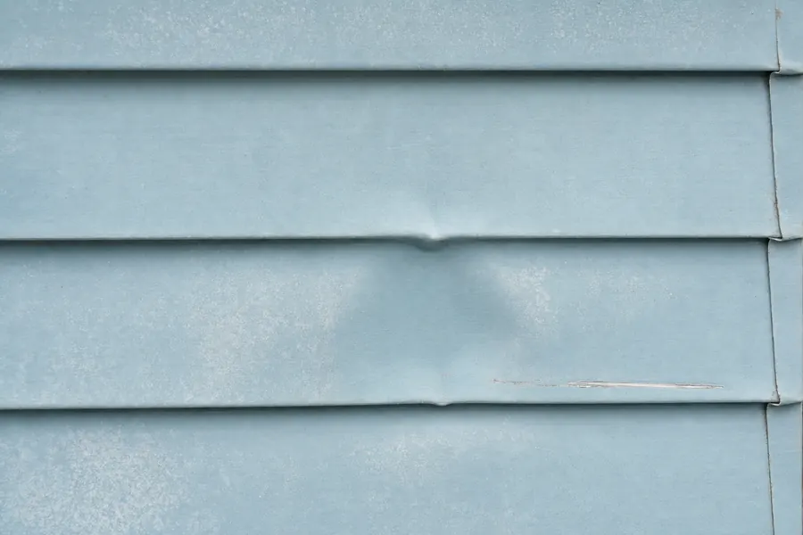 Painted aluminium lap siding in faded pale blue with a fist-sized permanent dent, chalky oxidised surface and a scratch showing bare metal.
